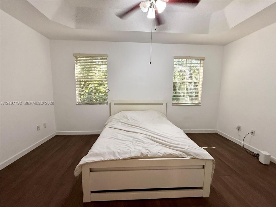Tray ceiling featuring a central multi-light ceiling fan, dual windows with horizontal blinds, and wood-finish flooring