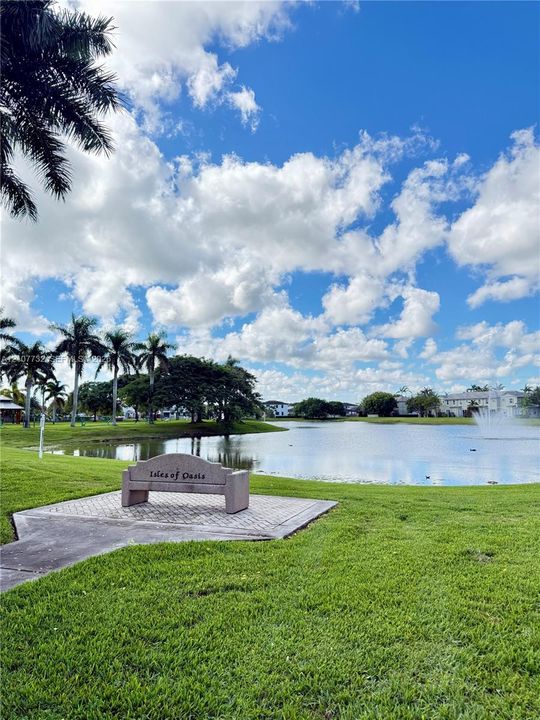 Manicured lawn featuring a stone bench on a paved platform, mature palm trees, and a water feature situated on a lake
