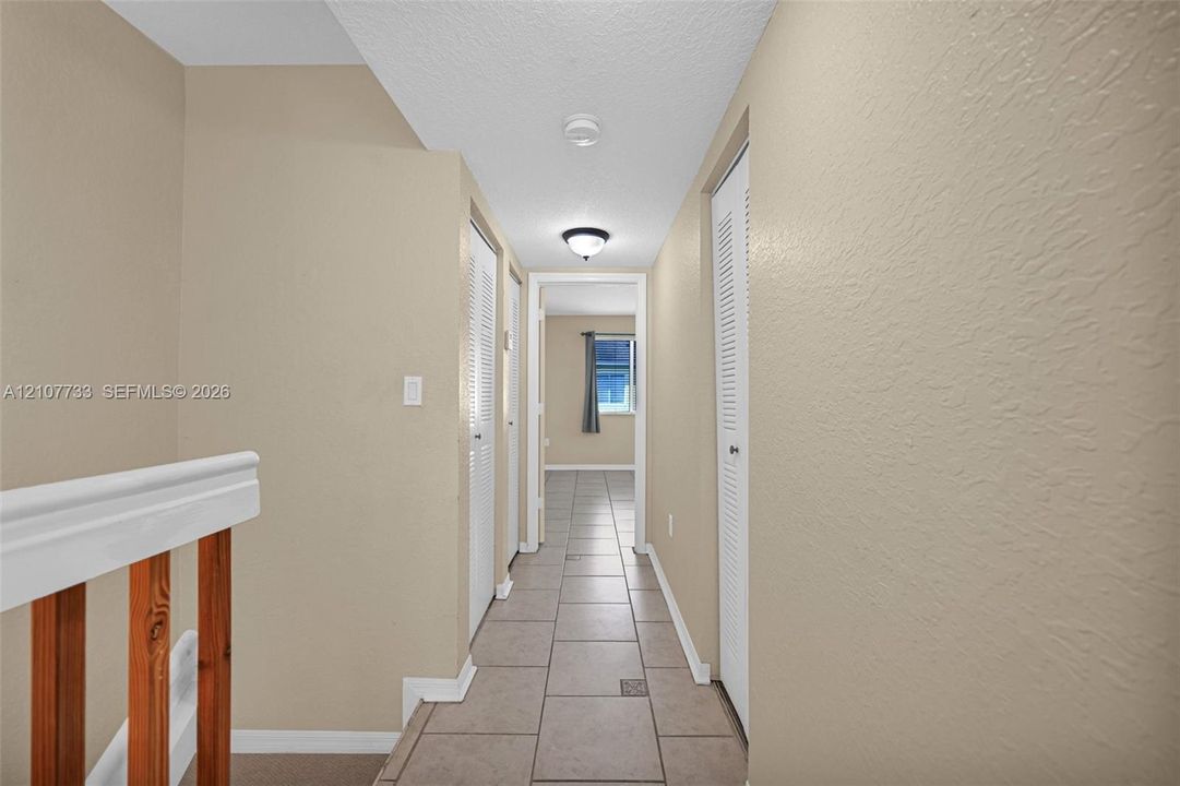 Hallway featuring large-format ceramic tile flooring, neutral wall tones, dual bi-fold closet doors, and wood-finish railing accents