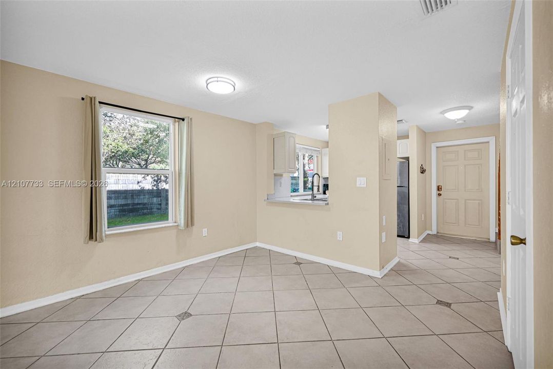 Tile flooring set on a diagonal pattern, neutral wall color, oversized window, and kitchen pass-through with a light-toned countertop