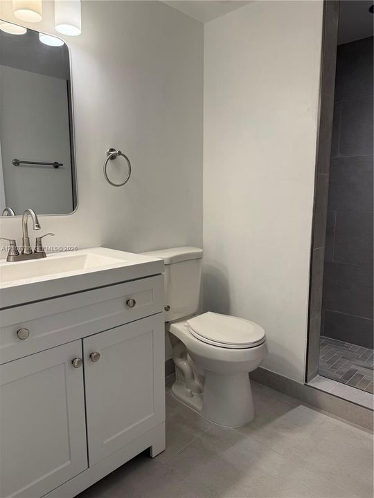 White vanity with integrated rectangular sink and metallic hardware, rounded rectangular mirror, three-light overhead fixture, and shower with dark tile surround and herringbone-patterned floor tile
