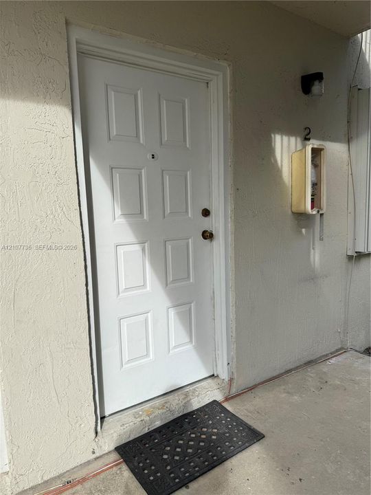 White six-panel entry door featuring a central peephole and brass-finish hardware, set within a stucco exterior wall