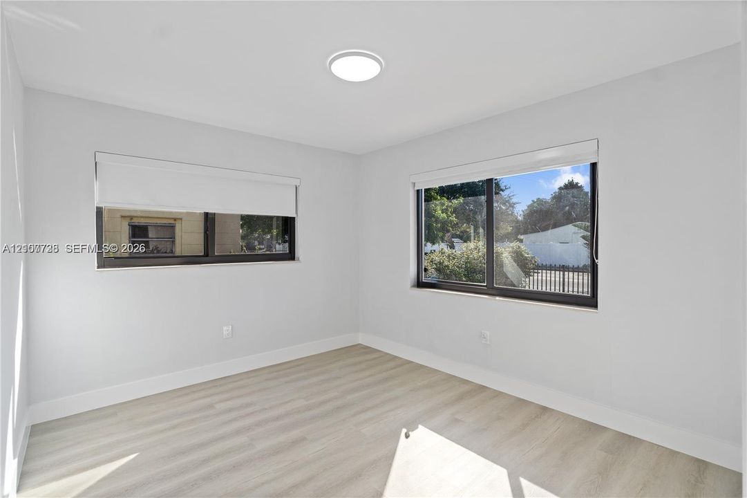 Neutral-toned interior space featuring wood-finish flooring, crisp white walls, dual windows with integrated shades, and a flush-mount ceiling light fixture