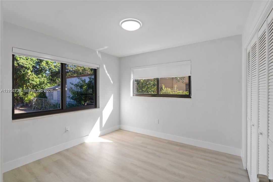 Neutral-toned room featuring wood-finish flooring, dual windows with roller shades, a flush-mount ceiling light, and floor-to-ceiling louvered-panel closet doors