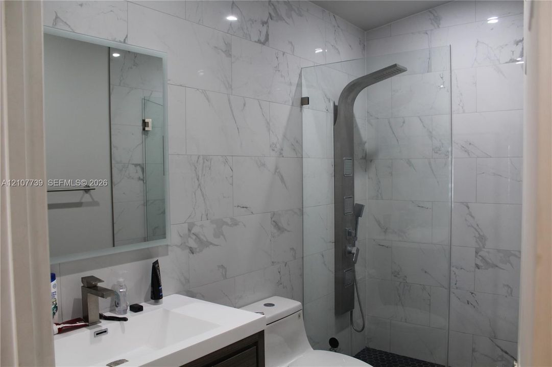 Bathroom featuring floor-to-ceiling stone-look wall tile, a walk-in shower with a glass panel and integrated shower panel system, an illuminated mirror, and a vanity with a white rectangular sink and metal faucet