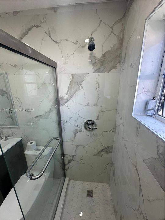Shower featuring large-format wall tiles with veining patterns, glass enclosure with metal hardware, polished chrome shower fixtures, and hexagonal floor tiling