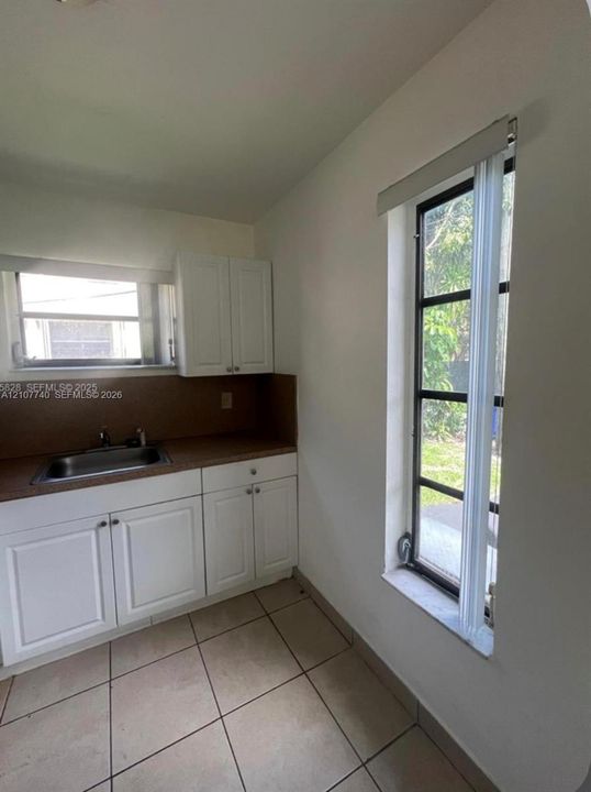 Kitchen area featuring white shaker-style cabinetry, solid-surface countertops, integrated stainless steel sink, large window with multi-pane grid, and light-toned tile flooring