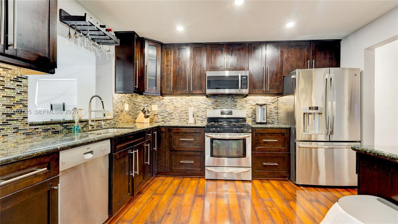 Kitchen featuring dark wood cabinetry, stainless steel appliances, multi-tonal stone tile backsplash, stone countertops, and wood-finish flooring