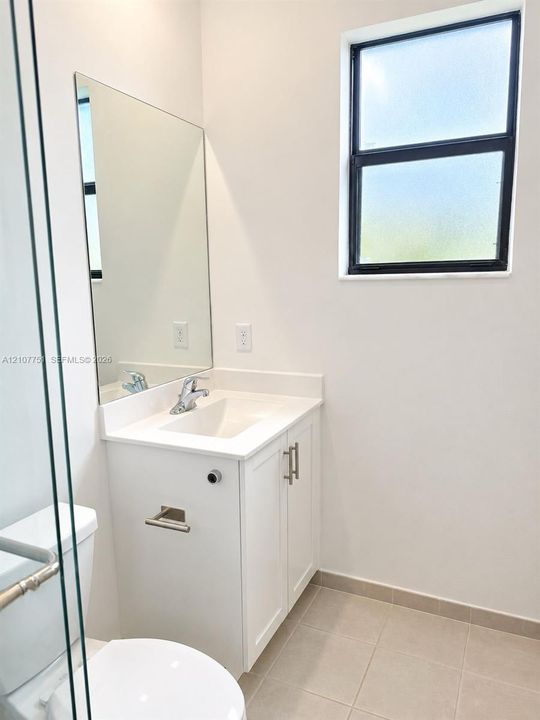 Bathroom featuring a white vanity cabinet with an integrated rectangular sink, solid-surface countertop, frameless wall-mounted mirror, and a window with a black frame