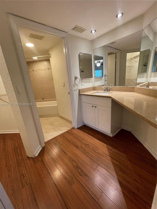 Bathroom featuring wood-finish flooring, white cabinetry with an extended vanity countertop, full-width wall mirror, and a tub-shower combination with tiled surround