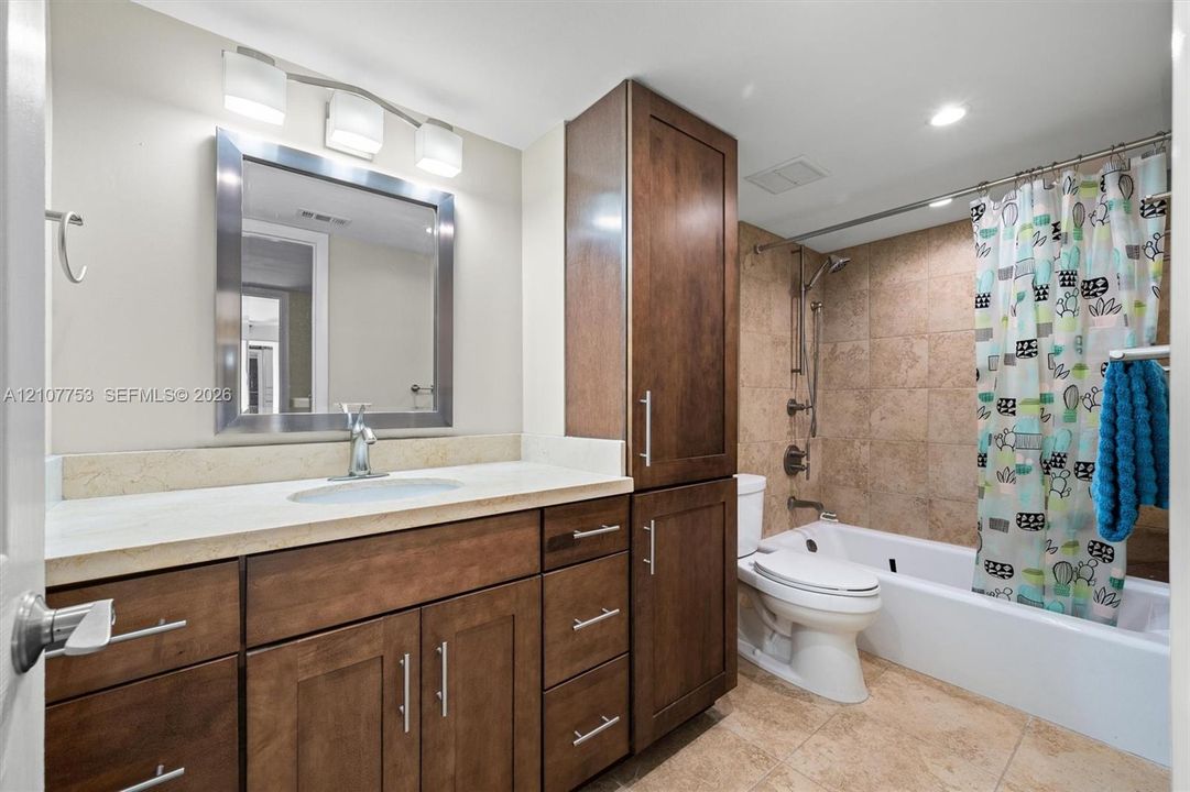 Bathroom featuring a vanity with dark wood-finish cabinetry, stone-composite countertop, full-height built-in linen tower, metallic wall-mounted vanity lighting, and a tub-shower combination with floor-to-ceiling tile surround