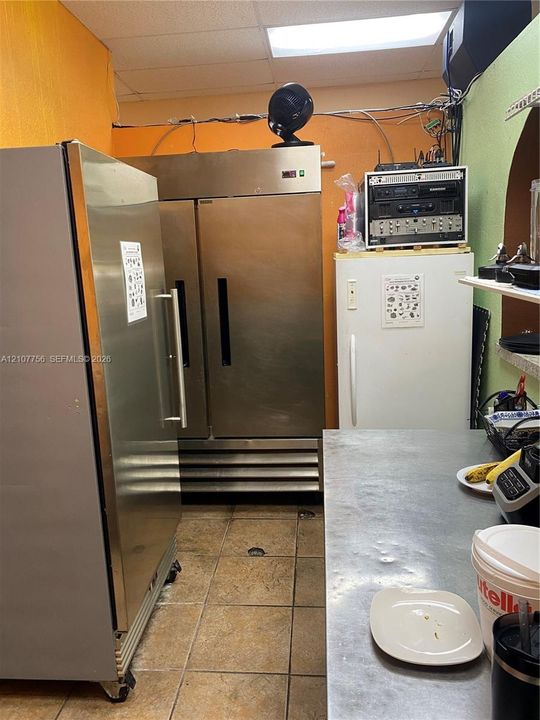 Commercial kitchen area featuring stainless steel dual-door refrigerator, upright refrigerator, stainless steel prep surface, and tile flooring