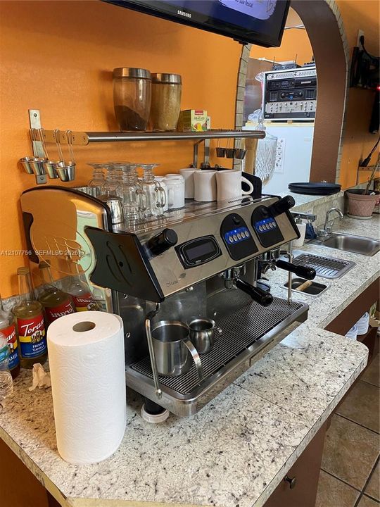 Commercial-grade espresso machine featuring stainless steel construction and digital interface, set within a preparation area with speckled stone-finish countertops, stainless steel sink, and wall-mounted metal shelving