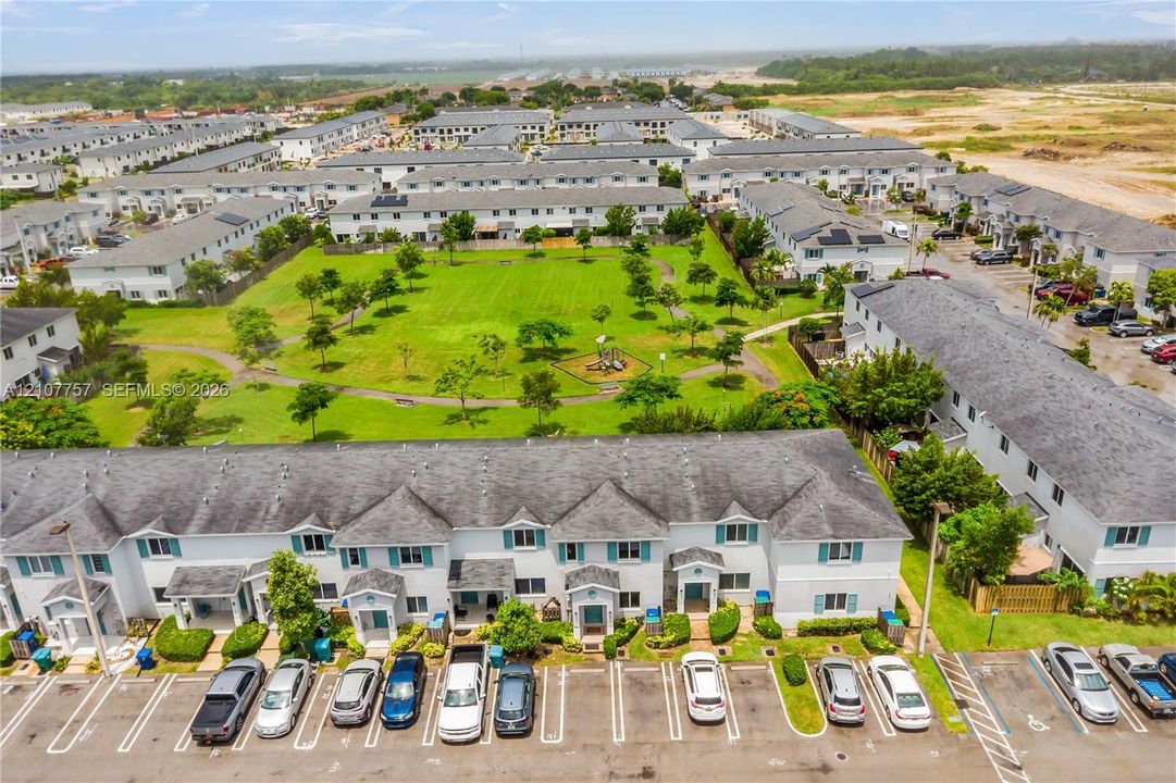 Aerial perspective of a multi-unit residential complex featuring light-toned siding, dark shingle roofs, designated surface parking, and extensive landscaped green space with paved pedestrian walkways