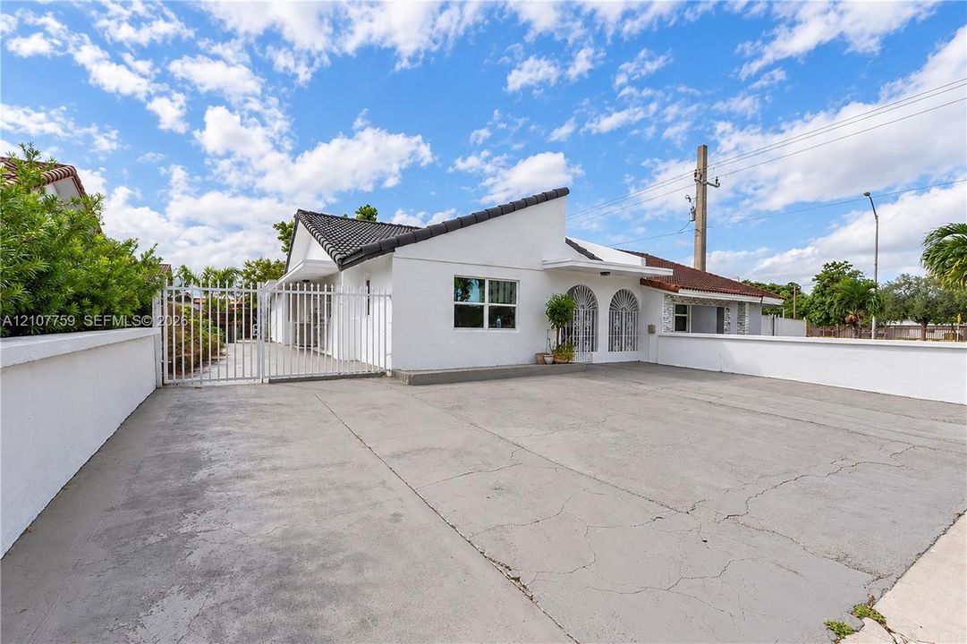 Single-story residence featuring a white stucco exterior, dark tile roof, arched entryway accents, and a gated concrete parking area enclosed by a perimeter wall