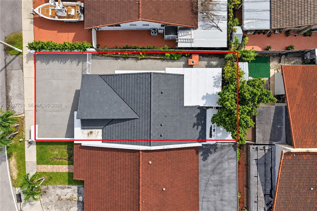 Aerial view of property featuring a dark-toned pitched roof, concrete driveway, covered patio area, and mature trees within the lot perimeter