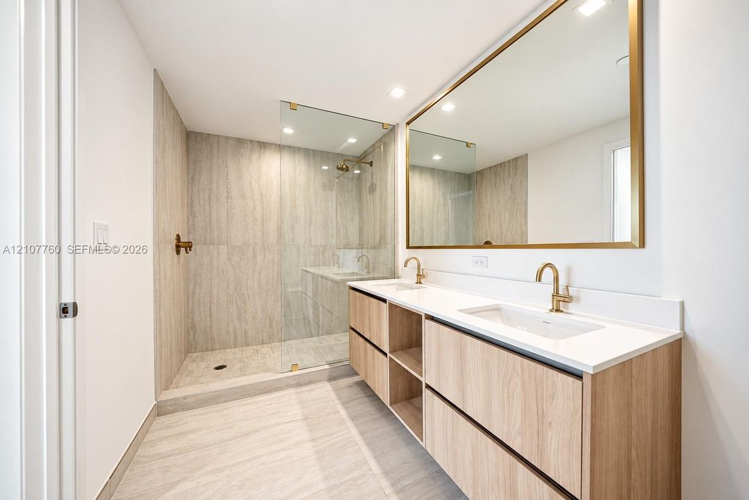 Wood-finish dual vanity with white countertop, integrated rectangular sinks, expansive gold-rimmed mirror, and walk-in shower with floor-to-ceiling stone-patterned tiling and gold-tone fixtures