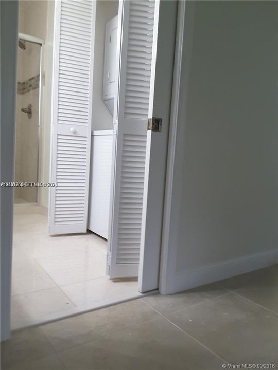 Laundry closet featuring white louvered doors and large-format tile flooring, accompanied by a bathroom enclosure with a glass shower door and decorative tile inlay