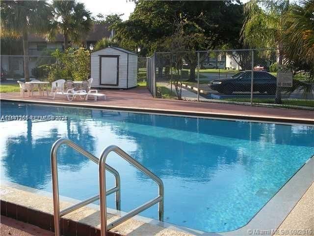 In-ground swimming pool with metal ladder entry, expansive paved patio, and detached storage shed surrounded by chain-link fencing