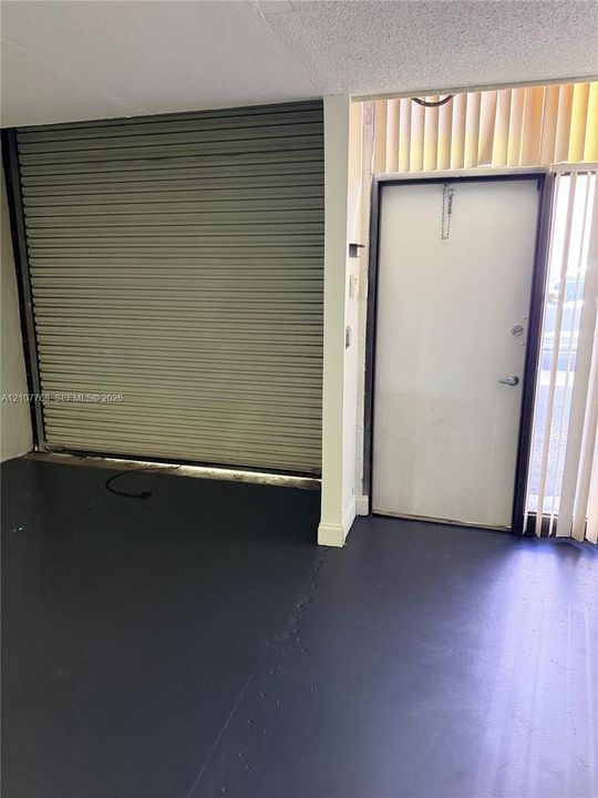 Interior space featuring a metal roll-down security shutter, solid-core entrance door, dark-toned flooring, and floor-to-ceiling vertical blinds