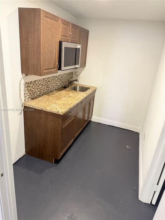 Kitchenette featuring wood-finish cabinetry, natural stone countertops, a mosaic tile backsplash, stainless steel microwave, and an integrated stainless steel sink