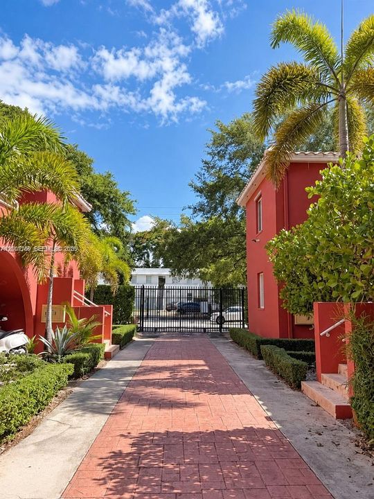 Red-toned stucco exterior featuring a paved walkway, mature palm trees, landscaped shrubbery, and gated entry