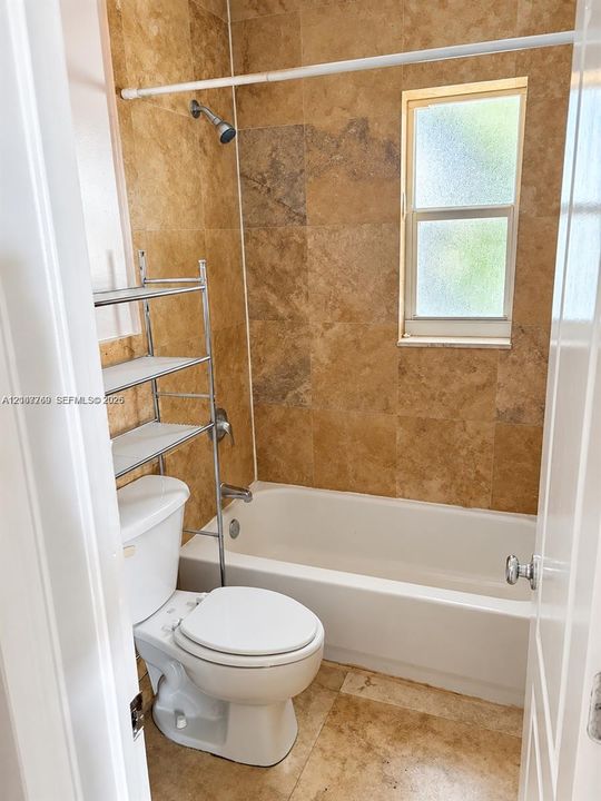Full-height natural stone wall tile in a warm finish, standard bathtub, and obscured-glass window