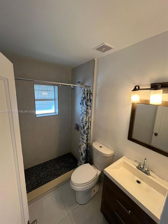 Bathroom featuring a wood-finish vanity with an integrated basin, wall-mounted mirror, neutral large-format tile flooring, and a walk-in shower with stone-style floor tiling