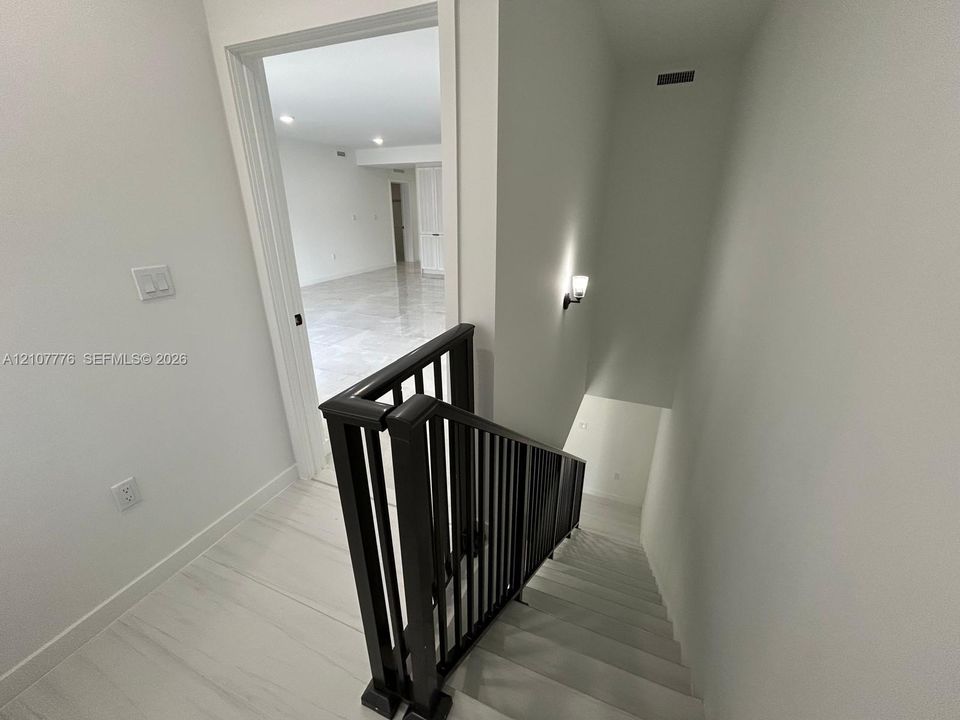 Staircase featuring a black metal balustrade with a wooden handrail, neutral flooring, wall-mounted light fixture, and tile flooring visible in the room beyond