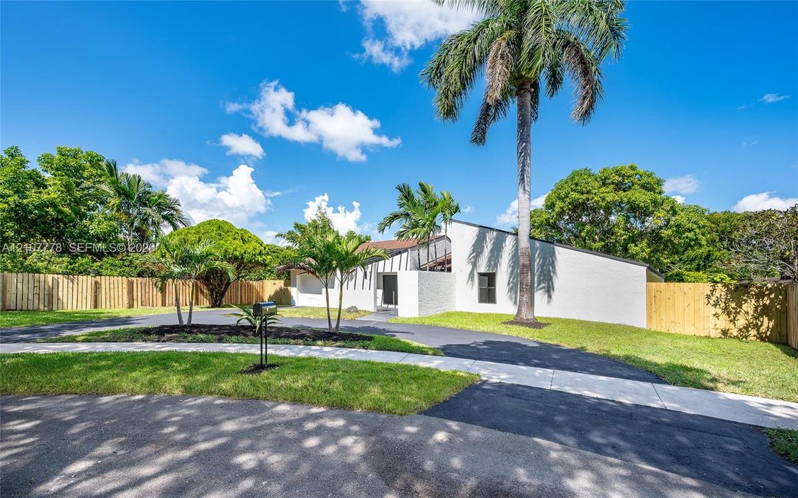 Exterior facade featuring white brick masonry, asphalt driveway, mature palm trees, and perimeter wood fencing