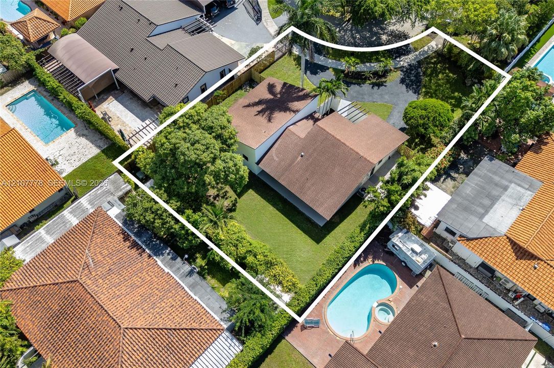 Aerial perspective of a property featuring a multi-section shingled roof, a manicured lawn, perimeter hedge fencing, and mature palm trees