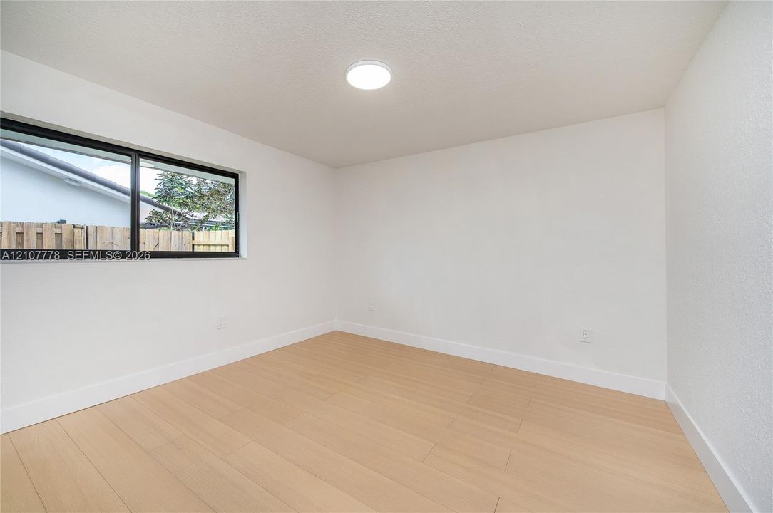 Neutral interior room featuring wood-finish flooring, crisp white walls, a single horizontal window with a black frame, and a flush-mount circular ceiling light