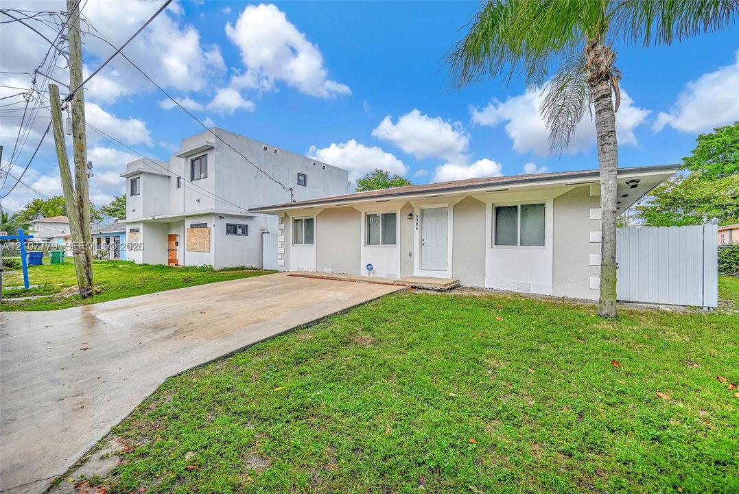 Single-story exterior featuring a light-toned stucco facade, concrete driveway, white vinyl privacy fencing, and a mature palm tree