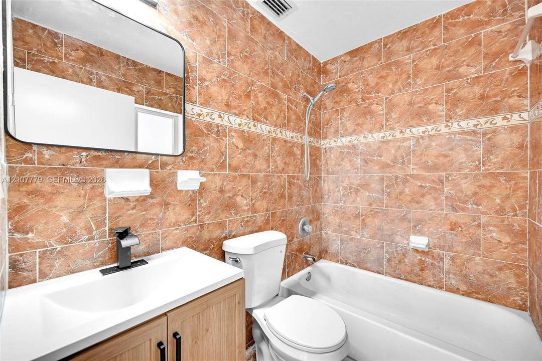 Bathroom featuring full-height ceramic tile walls with decorative inlay, a wood-finish vanity with an integrated countertop sink, matte black faucet hardware, and a frameless rectangular mirror