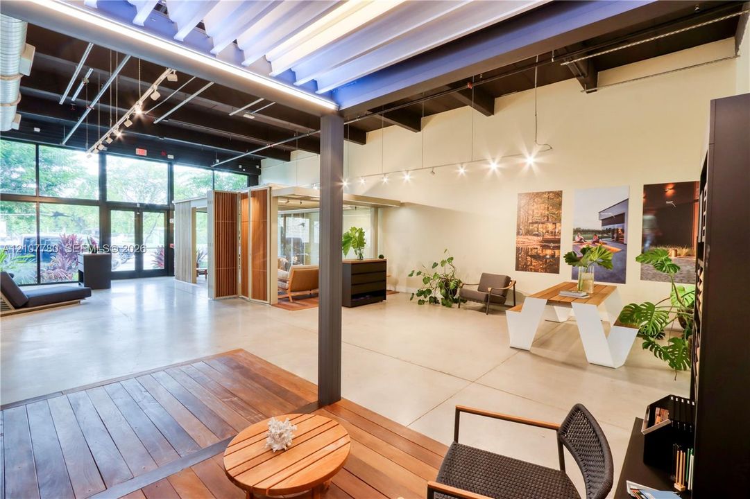 Open-concept interior featuring exposed structural steel beams, high ceilings with track lighting, floor-to-ceiling glass wall, polished concrete flooring, and an inlaid wood-finish platform
