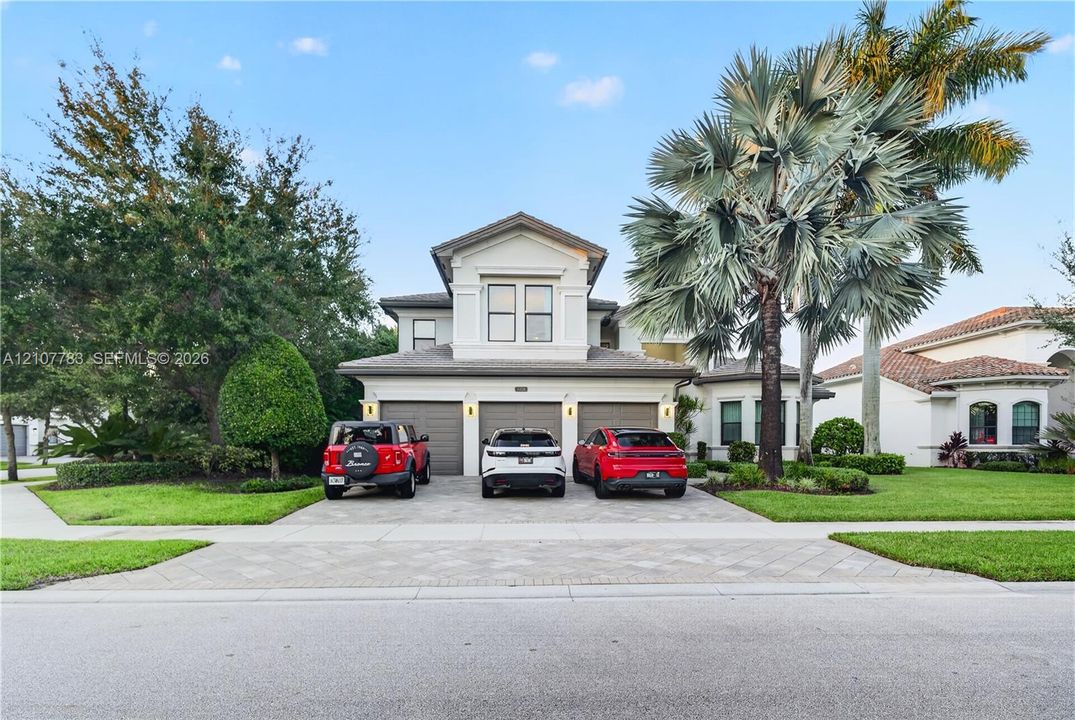 Two-story stucco exterior featuring a three-car garage, paver driveway, and mature palm landscaping