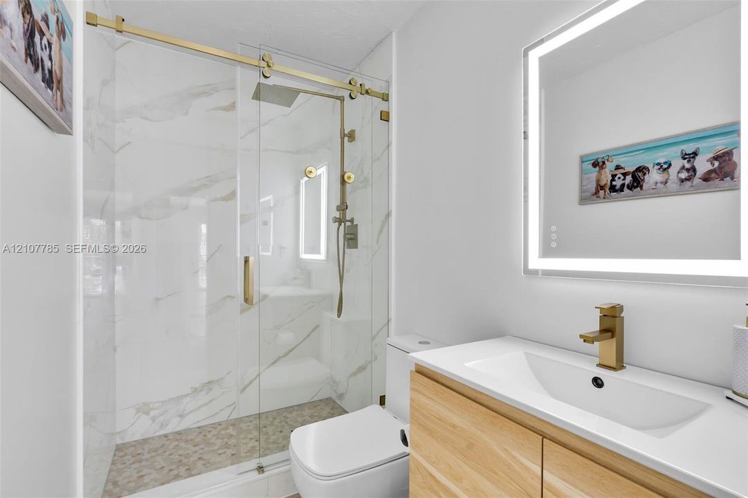 Walk-in shower with floor-to-ceiling stone-style tile, gold-finish sliding glass door, brass rain-style shower head, integrated LED-backlit mirror, and wood-finish floating vanity with vessel sink