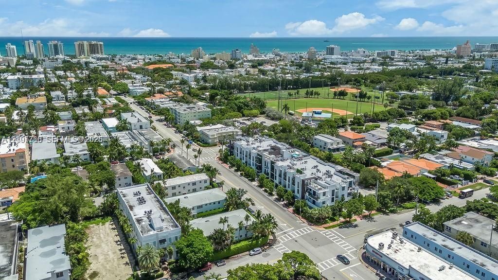 Aerial perspective of an urban neighborhood featuring coastal proximity, multi-story residential structures, mature tree canopy, and proximity to a community ball field