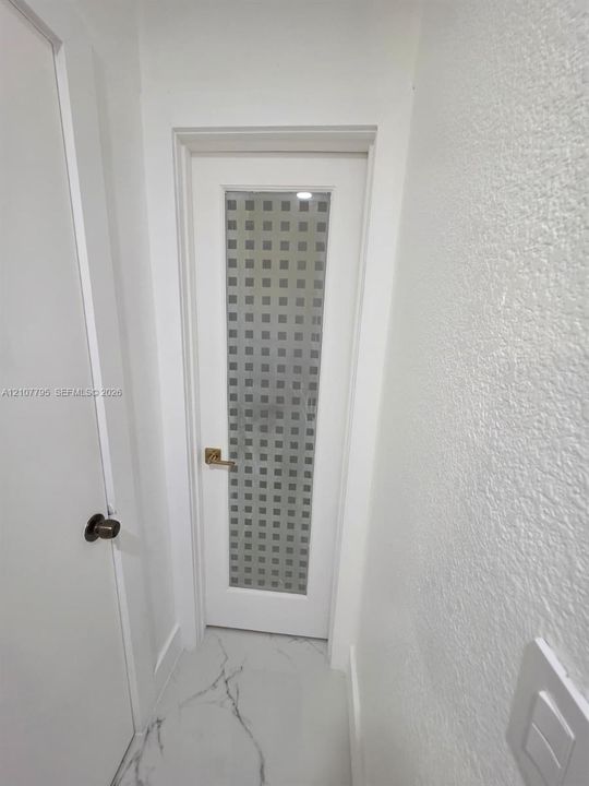Interior door featuring a white finish, frosted glass panel with a grid pattern, and metallic lever handle, set against white textured walls and white flooring with charcoal-toned veining