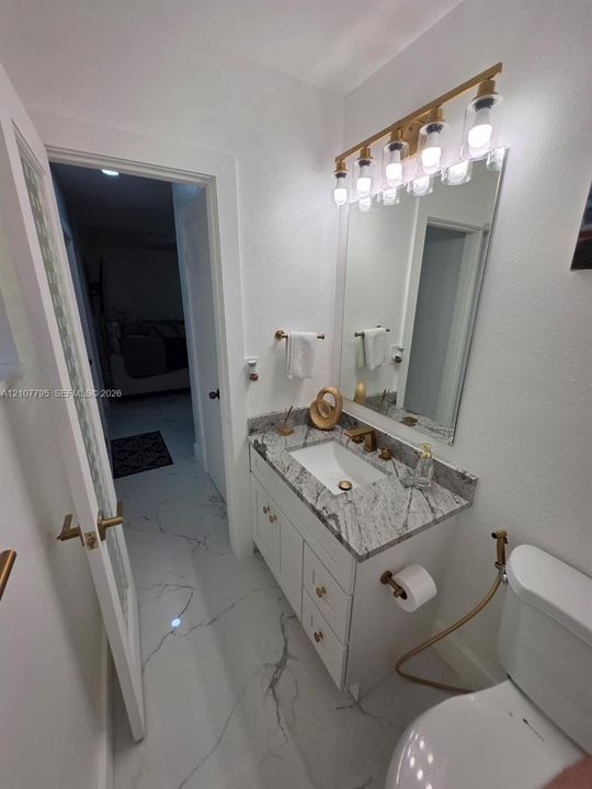 Bathroom featuring a white vanity cabinet with stone countertop, undermount sink, multi-bulb wall-mounted light fixture, gold-tone hardware, and large-format tile flooring