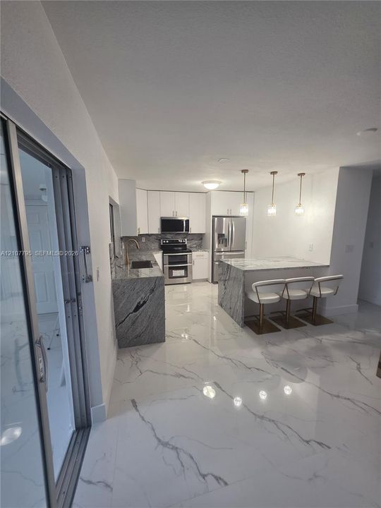 Kitchen featuring white shaker-style cabinetry, stone countertops with waterfall edge, stainless steel appliances, marble-patterned tile flooring, and three pendant lights
