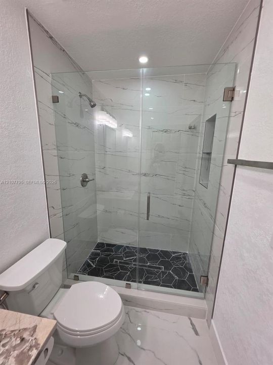 Walk-in shower featuring large-format stone-pattern wall tile, black hexagonal floor tile, frameless glass door, recessed wall niche, and metal shower fixtures