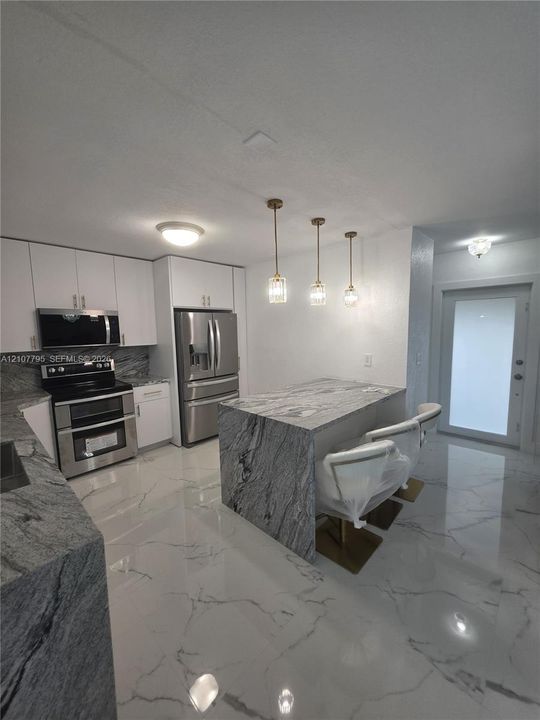 Kitchen featuring white shaker-style cabinetry, stone countertops with waterfall edge, stainless steel appliances, polished stone-finish floor tile, and three pendant light fixtures