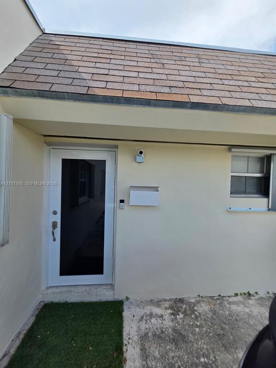 Exterior entry featuring a glass-panel door, shingle roof, mounted security camera, and wall-mounted mailbox