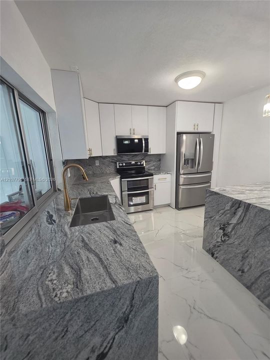 Kitchen featuring stone countertops, white cabinetry, large-format tile flooring, stainless steel appliances, and a gold-tone faucet