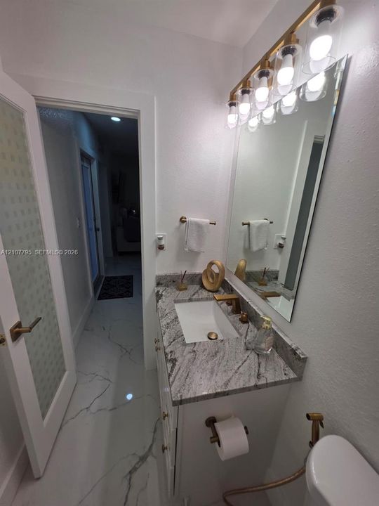Vanity featuring a stone countertop, rectangular undermount sink, and brass-finish hardware, paired with a multi-bulb vanity light, wall-mounted mirror, and tile flooring