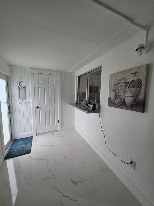 Interior space featuring large-format veined tile flooring, vertical plank wall paneling, a six-panel door, a window pass-through with stone-finish ledge, and a wall-mounted light fixture