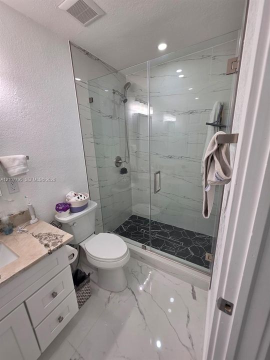 Walk-in shower featuring large-format stone-pattern wall tile, black geometric floor tile, and a frameless glass enclosure with brushed nickel fixtures, complemented by white cabinetry, patterned stone countertop, and white large-format flooring