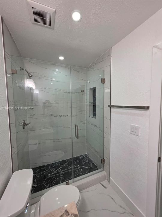 Walk-in shower featuring glass enclosure, wall-mounted shower head, built-in tiled niche, dark geometric floor tiling, and light-toned wall tile