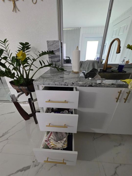 White cabinetry featuring matte gold-tone hardware, natural stone countertop, undermount stainless steel sink, bronze-finish high-arc faucet, and large-format porcelain tile flooring
